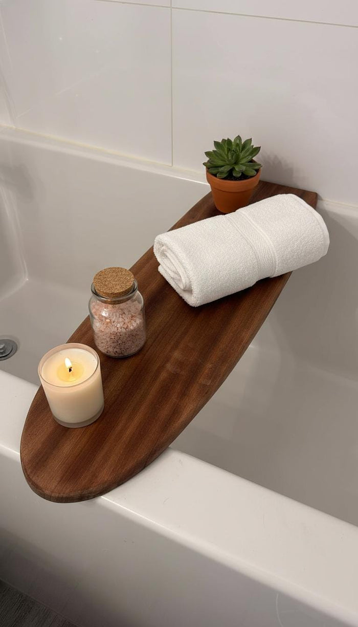 May include: A wooden bathtub tray holds a rolled white towel, a small potted succulent, a glass jar with pink bath salts, and a lit candle. The tray is dark brown and rests across the top of a white bathtub.