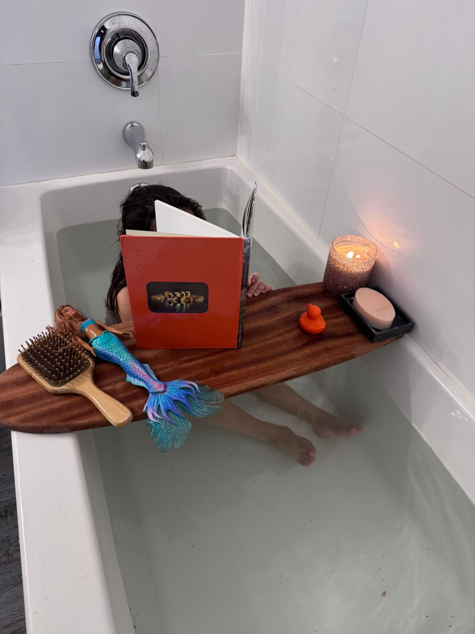 May include: A wooden bathtub tray holds a book, candle, soap, and a rubber duck. A mermaid doll and hairbrush rest on the tray. The person in the tub is reading a book with a red cover.