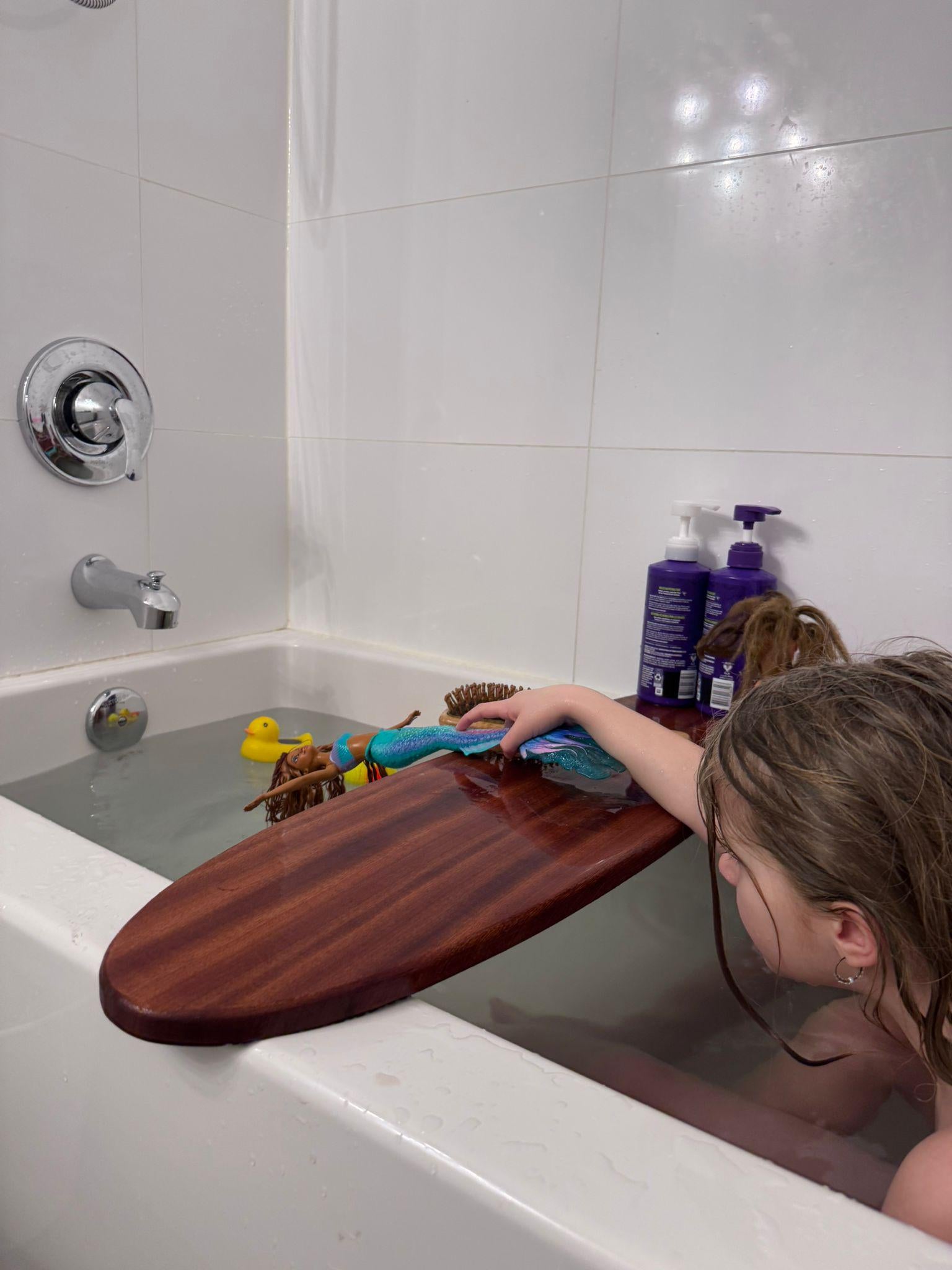 May include: A wooden bath tray with rounded edges, resting on the edge of a white bathtub. A child's hand is reaching for a mermaid doll and a hairbrush. A yellow rubber duck and other toys float in the water.