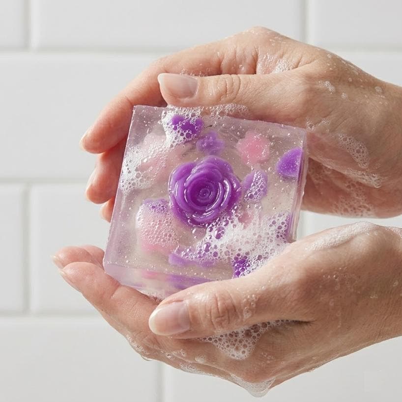 May include: A clear, square bar of soap with a purple rose embedded in the center. The soap also contains smaller purple and pink floral accents. The soap is surrounded by white suds.