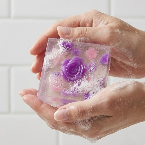 May include: A clear, square bar of soap with a purple rose embedded in the center. The soap also contains smaller purple and pink floral accents. The soap is surrounded by white suds.