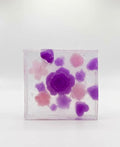 May include: A clear, square soap bar with embedded floral designs. The soap features a large, central purple rose surrounded by smaller purple, pink, and lavender flower and heart shapes. The soap is set against a white background.