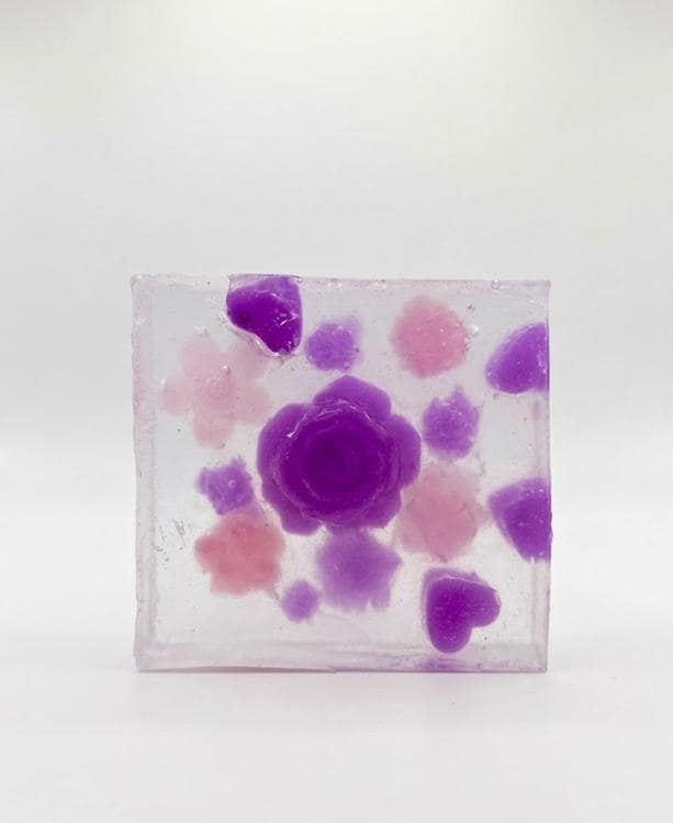 May include: A clear, square soap bar with embedded floral designs. The soap features a large, central purple rose surrounded by smaller purple, pink, and lavender flower and heart shapes. The soap is set against a white background.