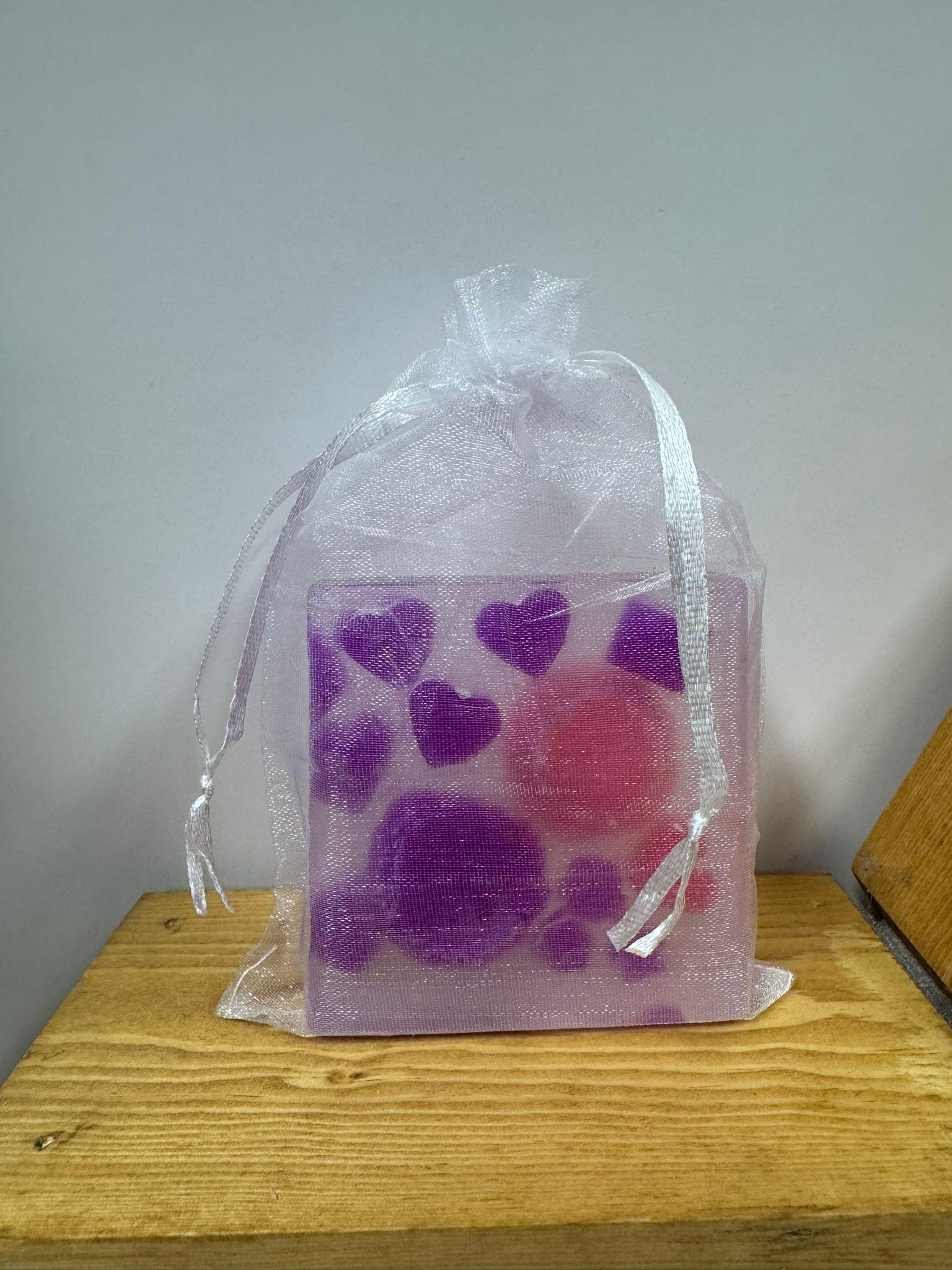 May include: A square soap bar with purple hearts and flower shapes, and a pink circle, is inside a sheer white drawstring bag. The soap is on a wooden surface.